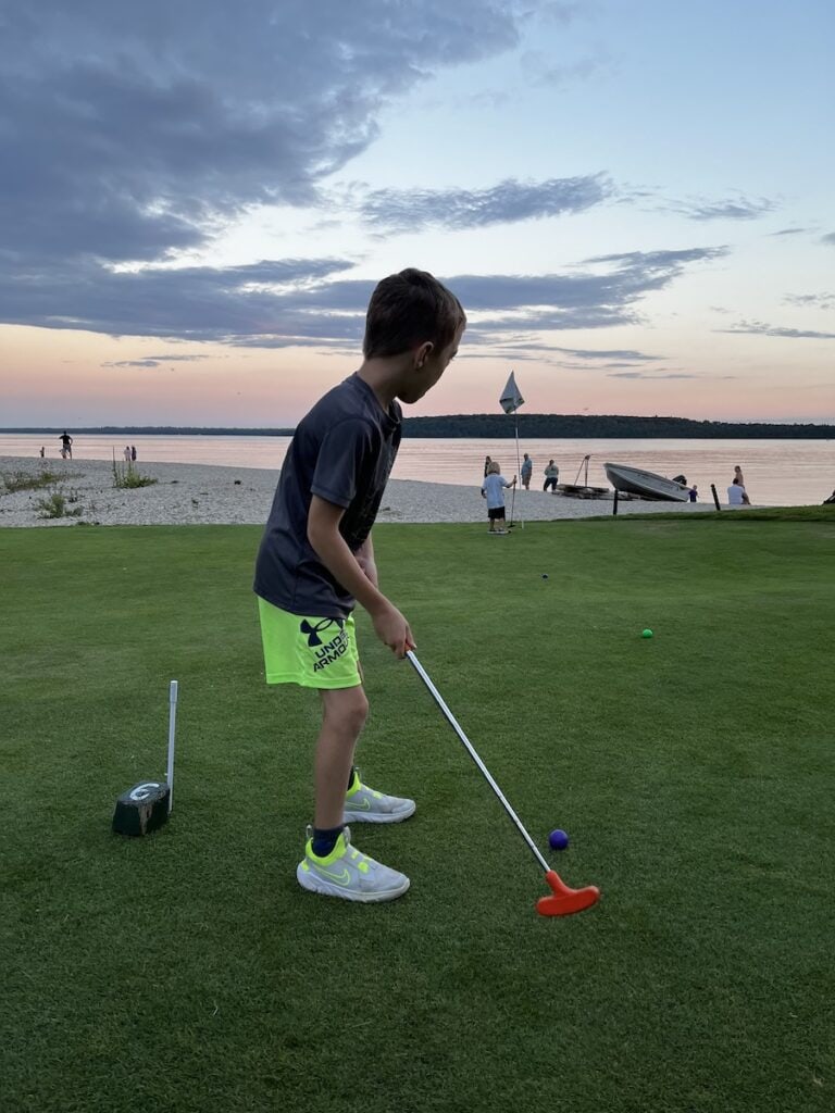 Boy ready to hit a golf ball with his golf club facing Lake Huron as the sun is setting on the miniature golf course at Mission Point Resort.