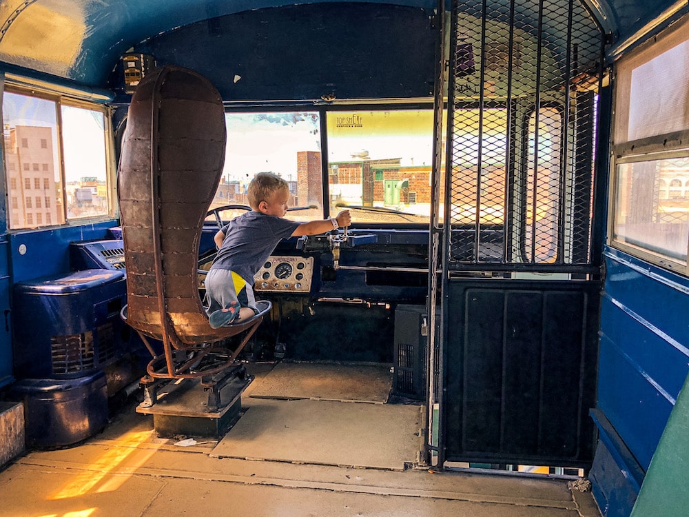 Child pretending to drive a school bus perched on the rooftop of the City Museum in St. Louis, with city buildings visible in the background.