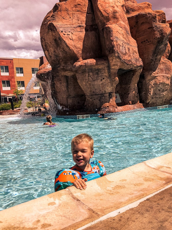 Young child in a colorful swim vest smiling at the edge of a resort-style pool with large red-rock formations and waterfalls at SpringHill Suites in Moab, Utah.