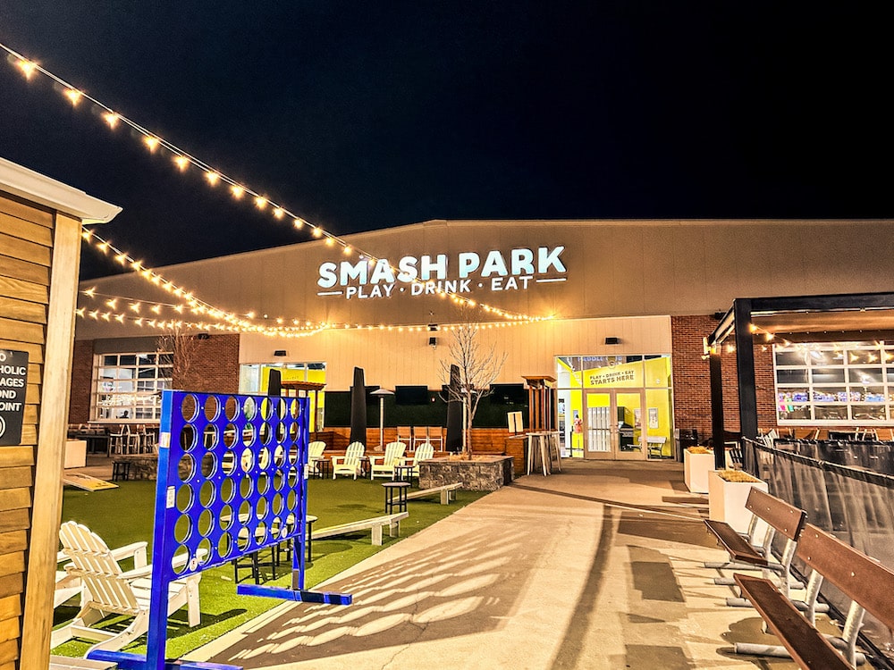 With the string lights on, the yard outside at Smash Park has cornhole, Giant Connect 4, and white adirondack chairs