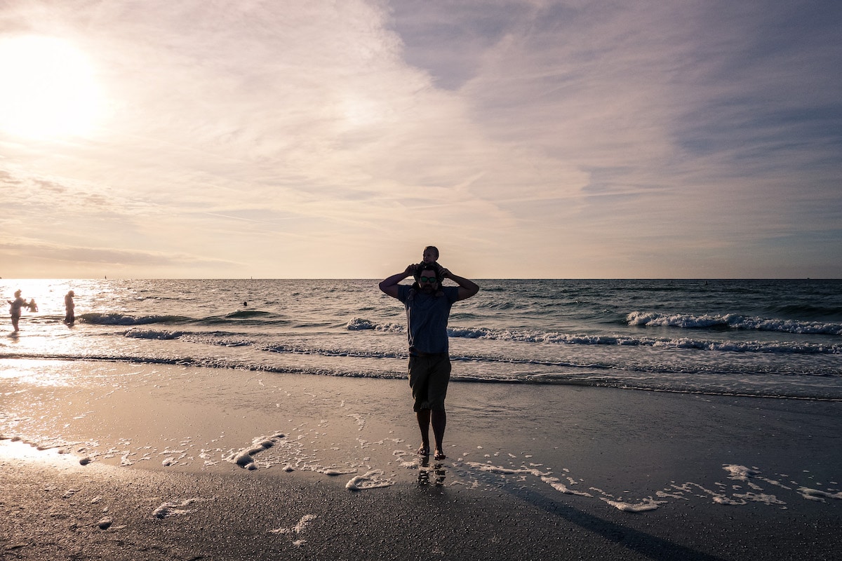 Father walking along the beach at sunset with a baby on his shoulders, waves rolling in behind them.