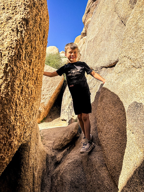 Boy standing between two humongous boulders taller than him in Joshua Tree National Park.