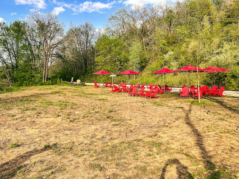Red adirondack chairs, side tables, and red umbrellas on the grass with lots of trees in the background