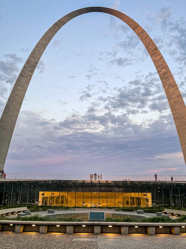 The Gateway Arch in St. Louis, Missouri against a pastel evening sky with soft clouds. Visitors walk along the base near the main entrance plaza, surrounded by curved walkways and modern landscaping.