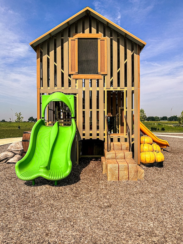 Side view of a playground shaped like a brown bar. It has a neon green double slide and stairs that look like hay bales.