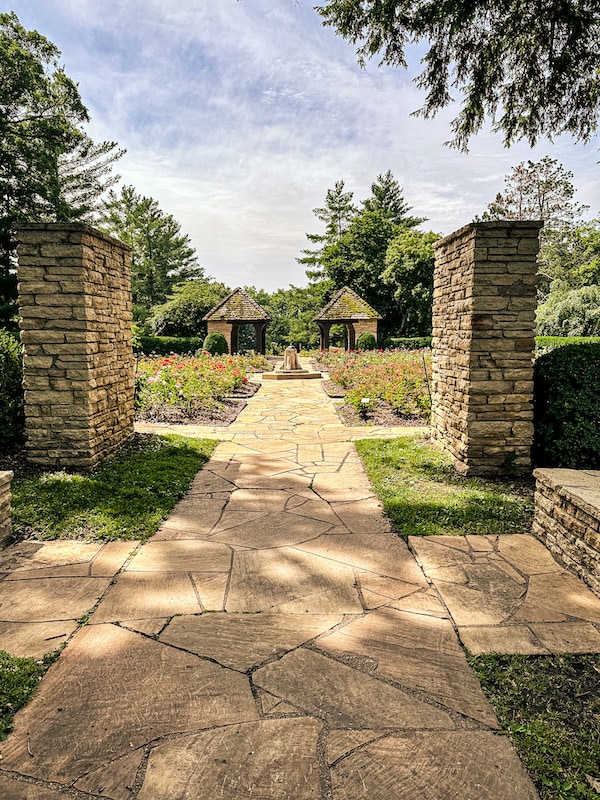 Stone walkway through the Clare and Miles Mills Rose Garden with two stone columns, roses, and two shelters in the background.