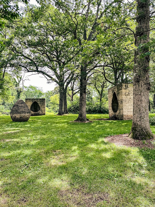 Abstract sculptures in in grassy field surrounded y trees.