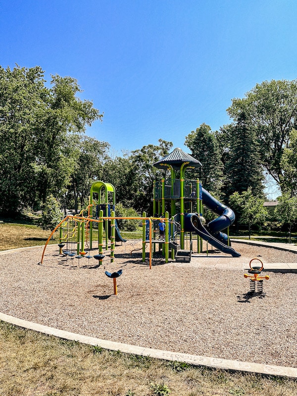 View of playground for older kids that includes a two level tower with a blue tunnel slide as well as an orange climbing structure to a green tower with a slide.