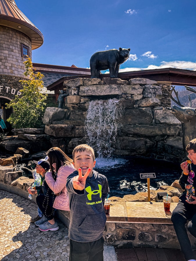 Boy holding up two fingers standing in front of a waterfall with a bear statue on the top of it