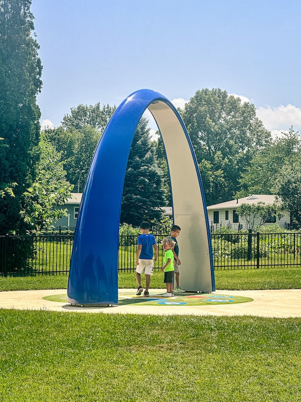 Three boys standing underneath the large blue arch interactive game at Wonder Spelen Park in Pella