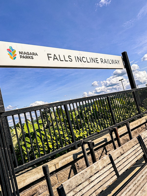 Sign for the Falls Incline Railway with a bench below it and a fence behind it.