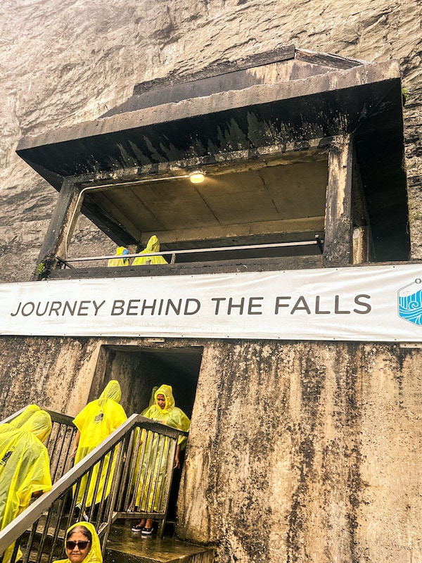Journey Behind the Falls sign with visitors wearing yellow ponchos walking back into the tunnel.