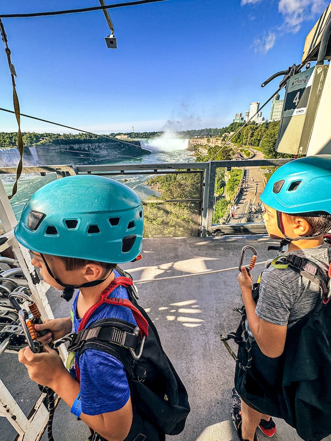 Two boys wearing teal helmets and harnesses waiting on the zipline platform with Horseshoe Falls in the background.