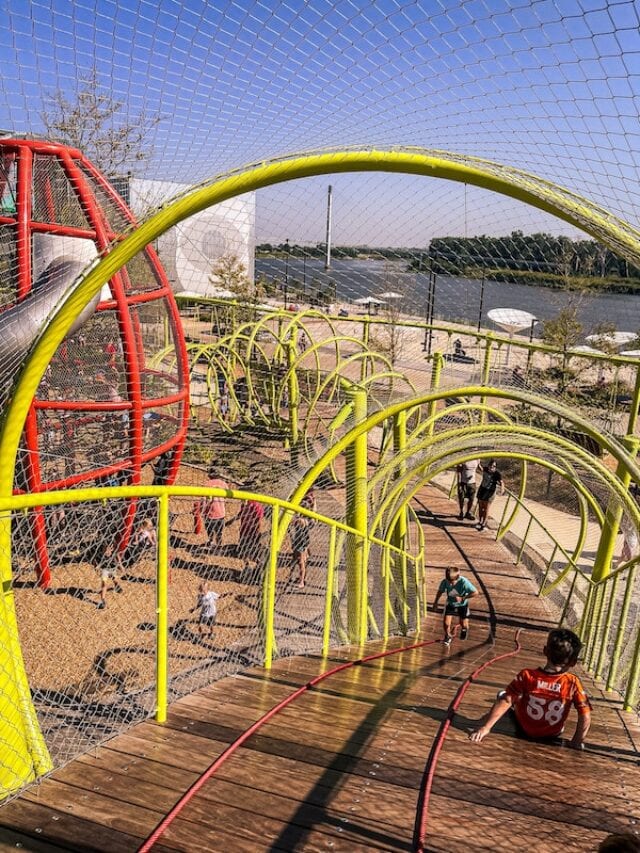Kids sliding down a wooden boardwalk play structure with the Missouri River in the background at the Discovery Playground in Omaha