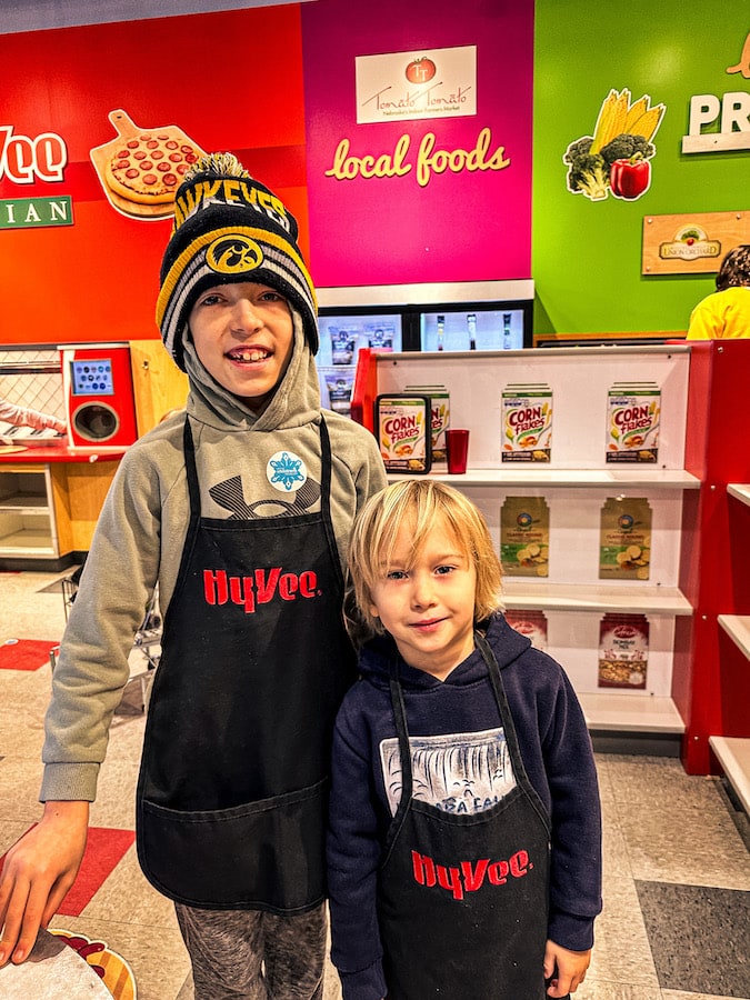 Two kids wearing HyVee aprons with the pretend grocery store behind them at the Omaha Children's Museum.
