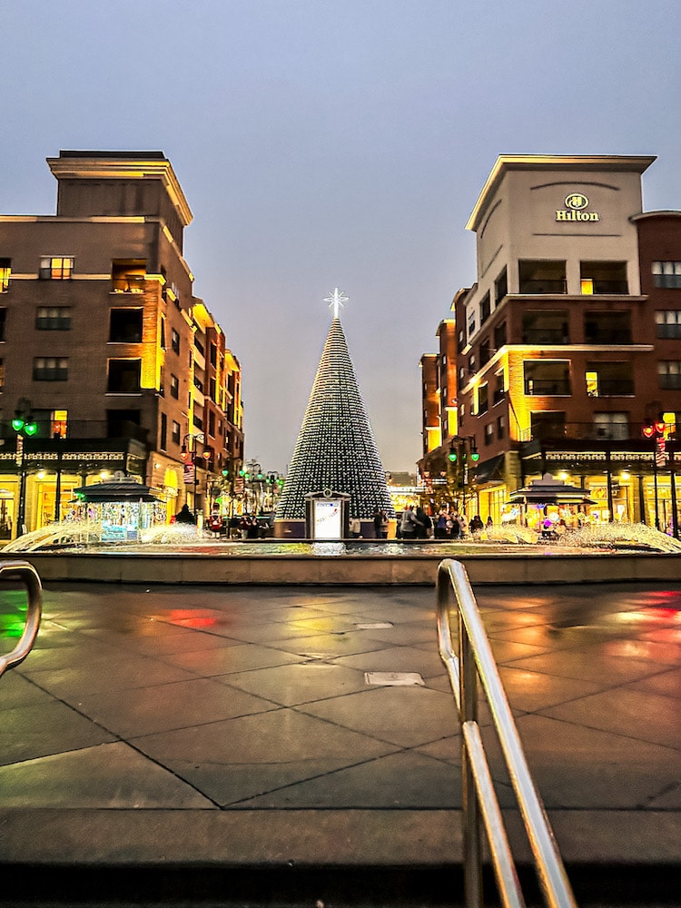 Large Christmas tree lit up at Branson Landing, with fountains in front and Hilton hotel buildings on each side at dusk.