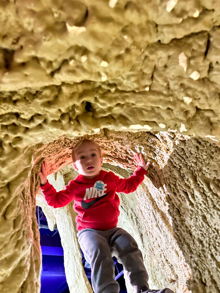 Young boy standing and holding on to the top of a cave inside of the Omaha Children's Museum.