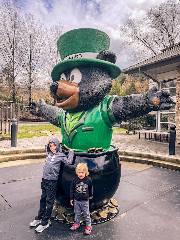 Two children posing with a large bear statue that is dressed like a leprachaun.
