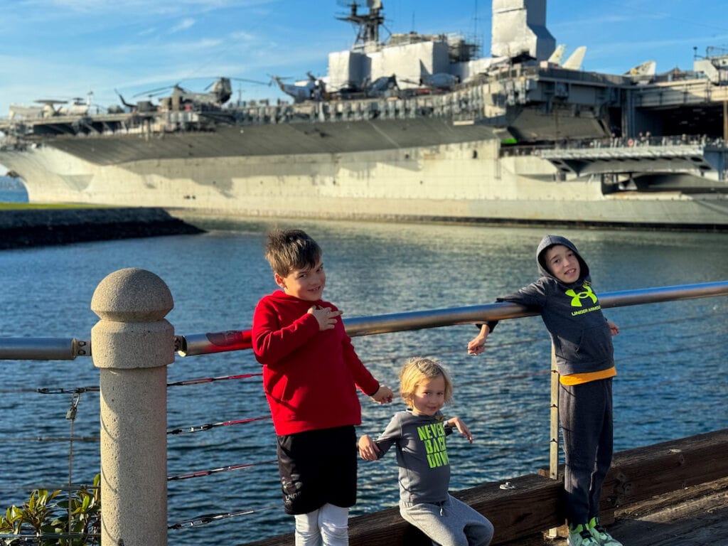 Three boys posing for a picture along a railing in front of the San Diego harbor and USS Midway aircraft carrier.