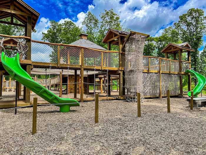 Treehouse Village playground at the Iowa Arboretum, featuring wooden towers, rope bridges, climbing nets, and bright green slides surrounded by trees.