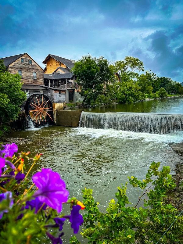 The Historical Old Mill in Forge, Tennessee with a view of colorful flowers in the foreground and the water wheel and log buildings in the back with a waterfall.