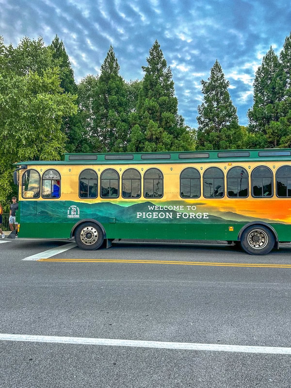 Green and yellow Pigeon Forge trolley at a stop with trees in the background.