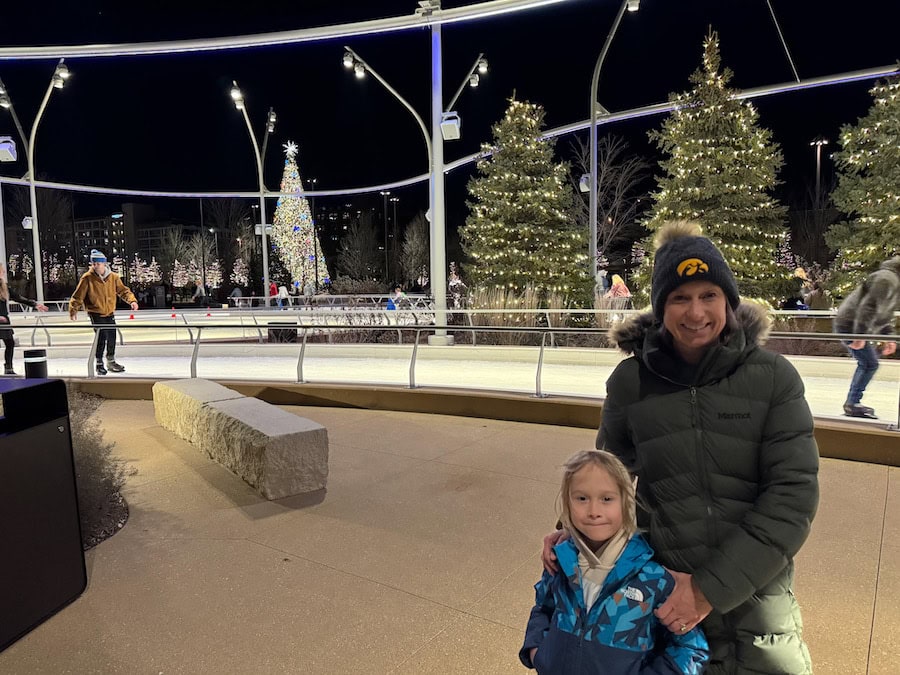Lady wearing a green winter coat and stocking hat with a young boy with a blue winter coat standing next to the Skate Ribbon in Heartland of America Park with the decorate evergreens and large Christmas tree in the background.