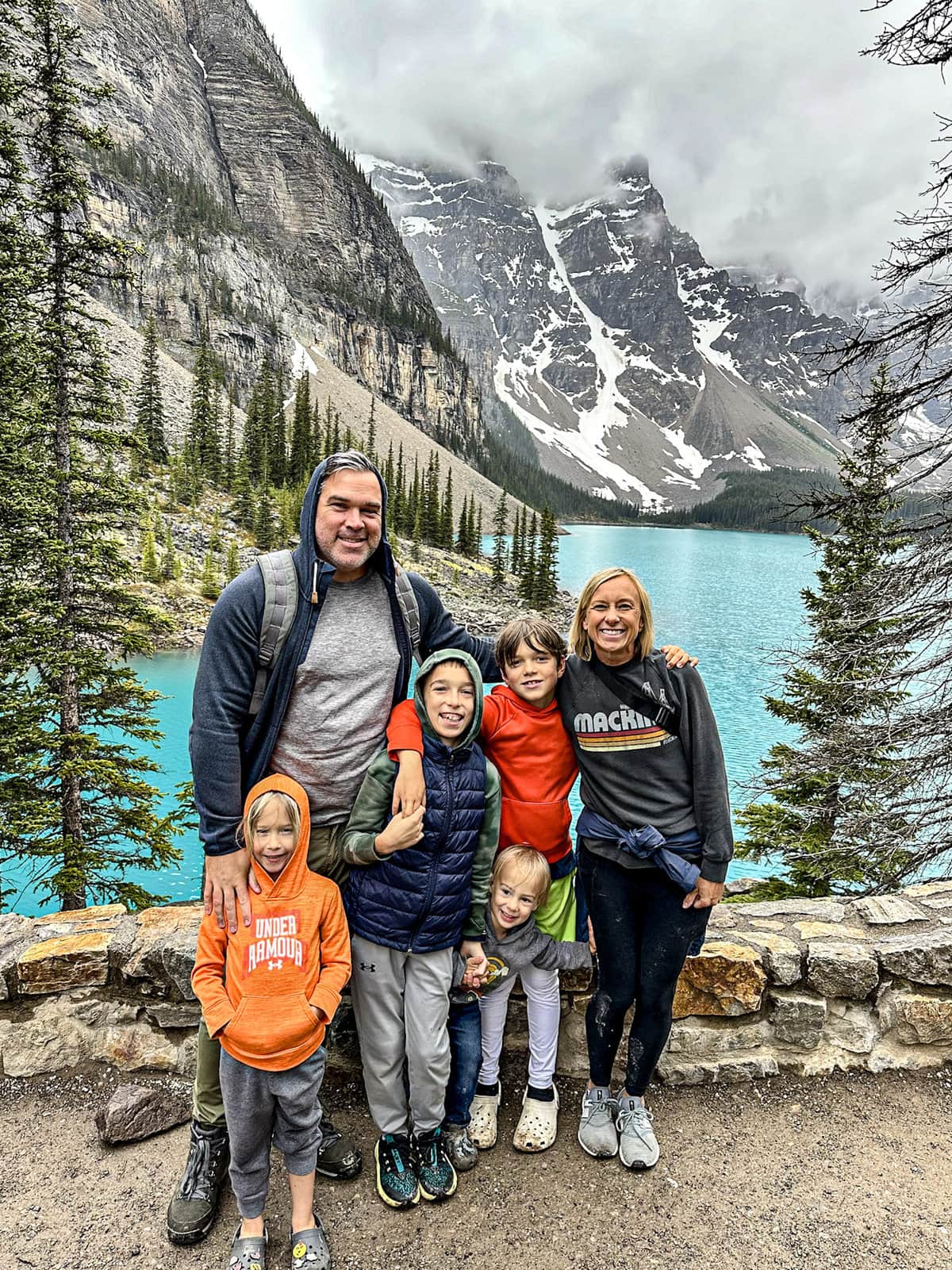 Family of six posing with the turquoise waters of Lake Moraine and snowy mountains in the background in Banff National Park.