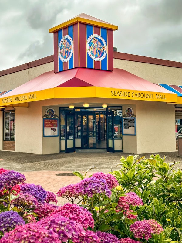 Entrance to the Seaside Carousel Mall in Seaside, Oregon, with colorful signage and a bright yellow and red awning, surrounded by blooming purple and pink hydrangeas.