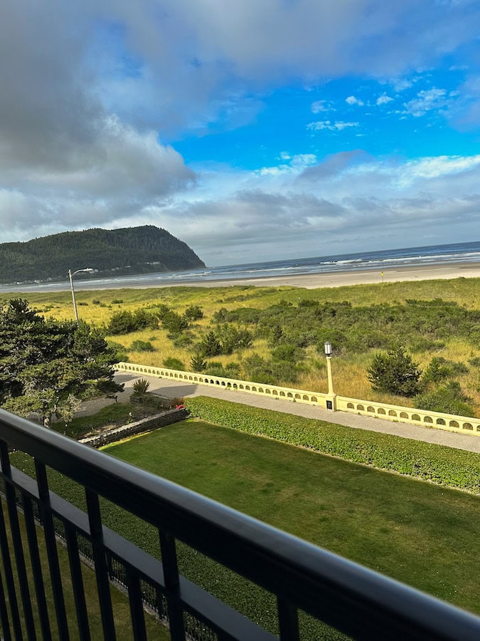 Ocean view from a Seaside, Oregon condo balcony, overlooking the Promenade, grassy dunes, and sandy beach with Tillamook Head in the distance under a partly cloudy sky.





