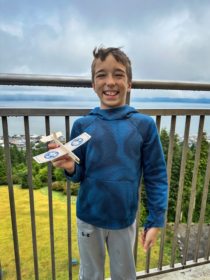 Smiling boy holding a wooden glider at the top of the Astoria Column, with scenic views of Astoria and the Columbia River in the background.


