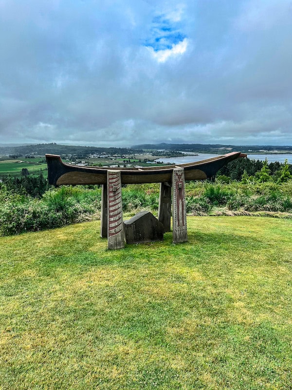 Wooden canoe sculpture on display near the Astoria Column, overlooking the Columbia River and surrounding green landscape on a cloudy day.