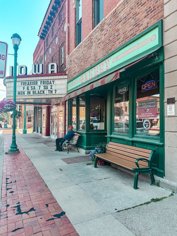 Street view of The Bakery Unlimited in downtown Winterset, Iowa, with its green storefront, large bakery sign, and a wooden bench out front, located next to the historic Iowa Theater marquee.