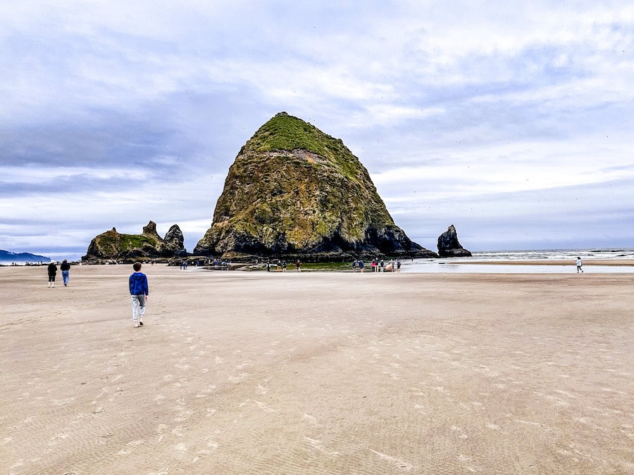 People walking across the wide sandy beach toward Haystack Rock in Cannon Beach, Oregon, on an overcast day.