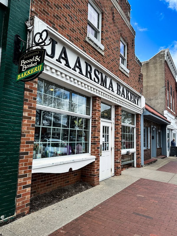 Front exterior of Jaarsma Bakery in Pella, Iowa, with its red brick facade, large white-framed windows, and hanging green and yellow Dutch-style bakery sign.