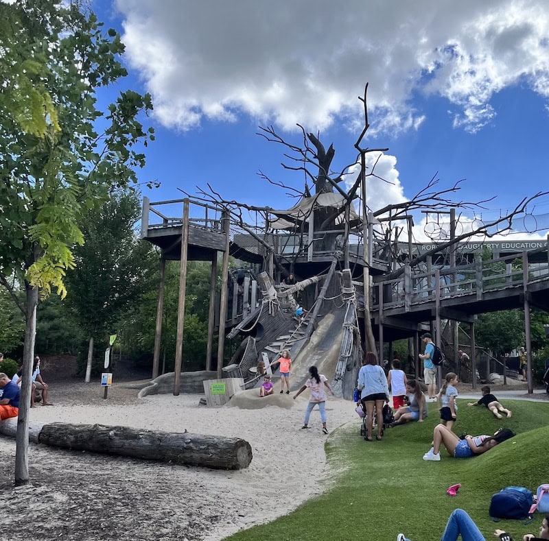 Children play on a large wooden treehouse-style playground with slides and climbing features in the Bay Family Children’s Adventure Trails at Omaha’s Henry Doorly Zoo.