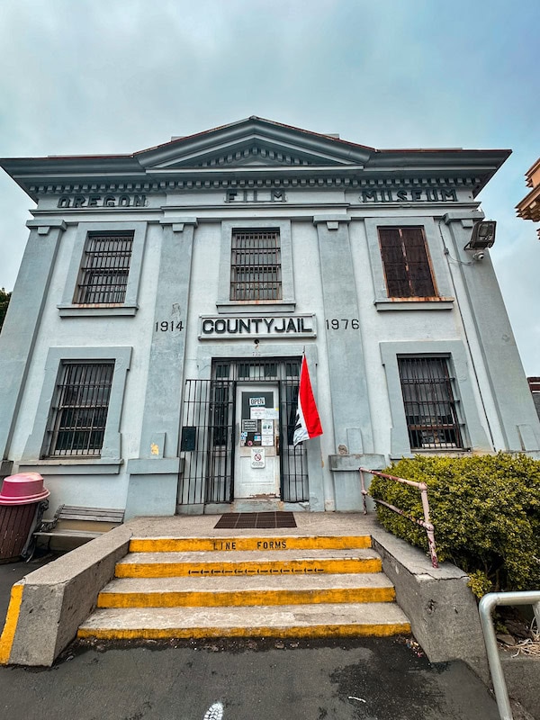 Exterior of the Oregon Film Museum, located in the historic Clatsop County Jail building in Astoria, Oregon, with barred windows and a red flag by the entrance.