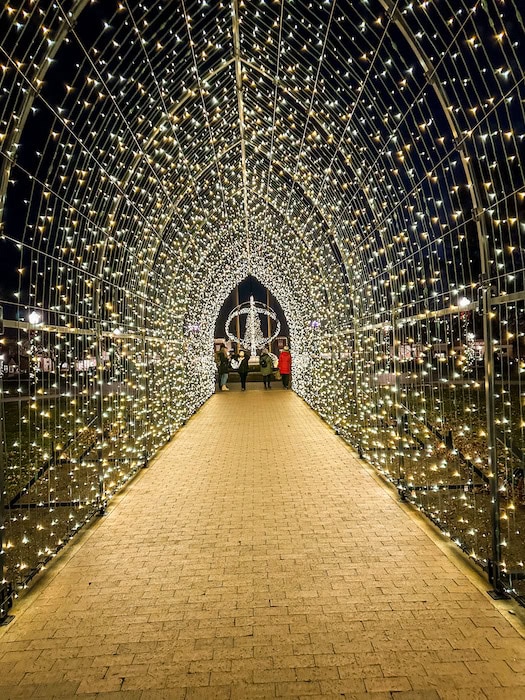 Illuminated light tunnel at night in Pella, Iowa, with thousands of twinkling white lights forming an arched walkway. People stand at the far end of the tunnel, with the Pella sundial lit up in the background as part of the holiday display.