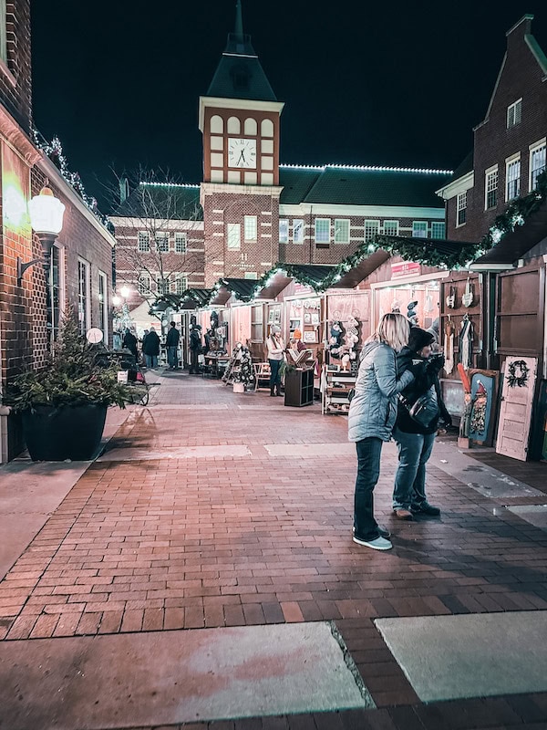 Evening scene at Kerstmarkt, Pella’s Dutch Christmas Market, with people browsing festive wooden vendor stalls decorated with garlands and lights. The brick-paved walkway leads toward a tall clock tower in the background.