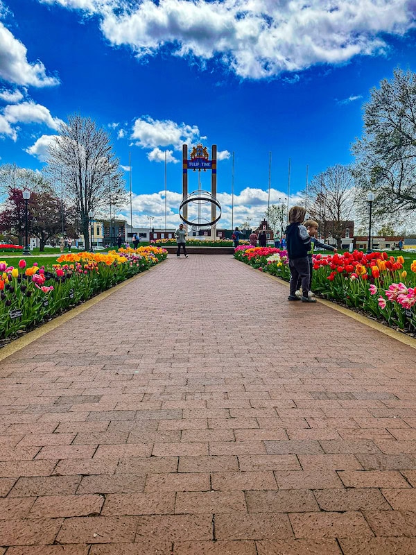 Brick pathway lined with colorful tulips in full bloom leading to the Tulip Time monument in Pella, Iowa. Two children stand near the flowers on the right, with people walking toward the monument under a bright blue sky with scattered clouds.