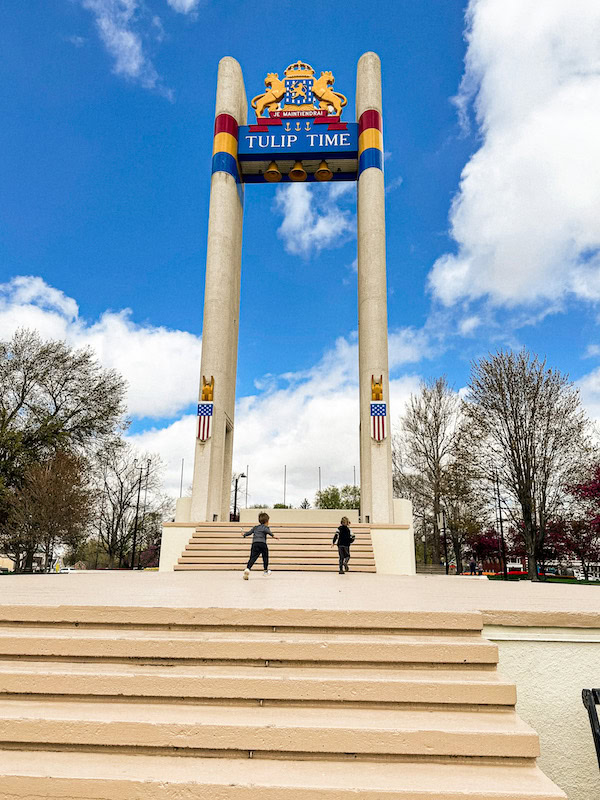 Two children run up the steps toward the tall Tulip Time monument in Pella, Iowa, featuring two vertical columns topped with the city crest and “Tulip Time” sign. Trees and a partly cloudy blue sky frame the background.