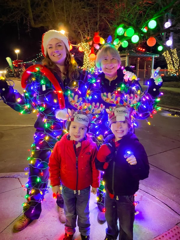 Kids posing with two ladies covered in Christmas lights at Jingle in the Junction in Valley Junction, Des Moines, surrounded by festive holiday decorations.