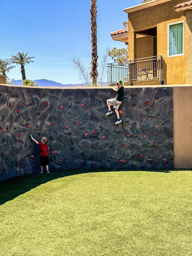 Two boys bouldering on a an outside rock wall with turf in front of it