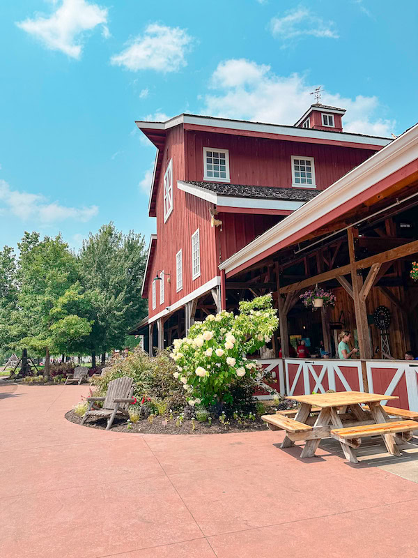 Scenic view of the red barn-style entrance building at Deanna Rose Children’s Farmstead in Overland Park, Kansas, with picnic tables, blooming flowers, and families enjoying the sunny day at this popular family attraction.
