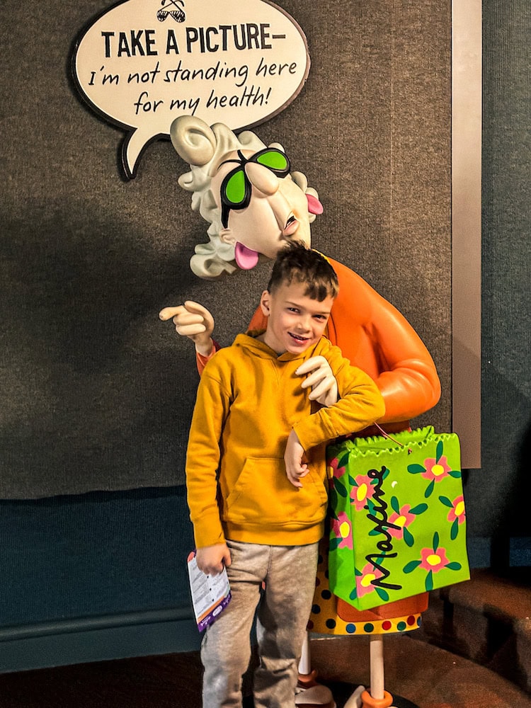 Young boy smiling in front of the Maxine statue at the Hallmark Visitors Center in Kansas City, posing next to the humorous sign that says, “Take a picture—I’m not standing here for my health!”