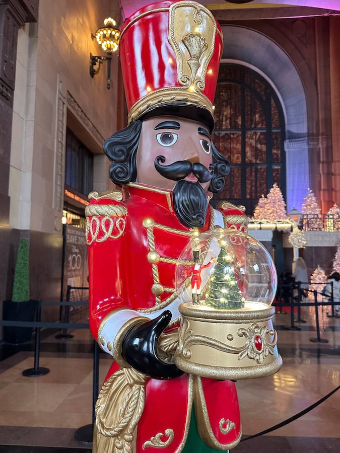 Festive nutcracker statue holding a glowing snow globe on display inside Union Station in Kansas City, surrounded by twinkling Christmas trees and lights.