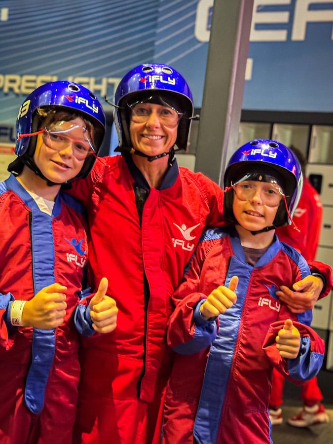 Mom and two young boys wearing red and blue iFLY flight suits and helmets, smiling and giving thumbs-up before their indoor skydiving experience at iFLY Kansas City.