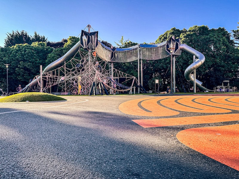 The large climbing structure at the Artists at Play Playground in Seattle, featuring tall rope nets, enclosed slides, and elevated walkways with trees and open play space surrounding it.