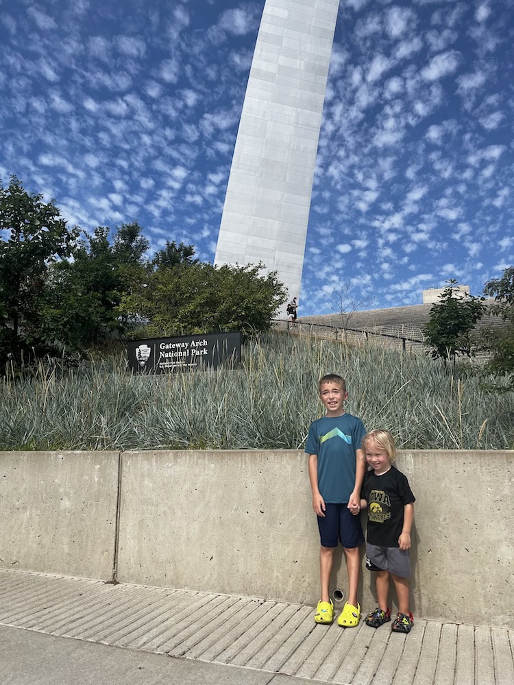 Two children standing in front of the Gateway Arch National Park sign with the Arch rising behind them on a partly cloudy day.