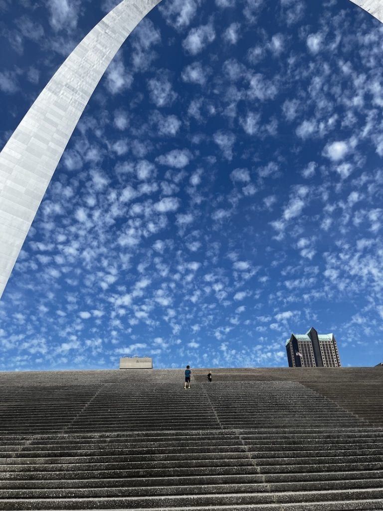 Two children climb the long concrete steps beneath the Gateway Arch in St. Louis, with a blue sky filled with scattered clouds overhead.